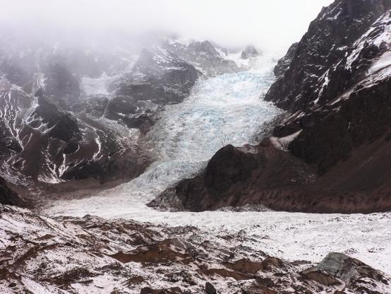 In the photo, you can see a large mountain of black and brown rocks. A white and blue glacier of eternal snow descends the mountain. Part of the peaks are covered in snow and fog.