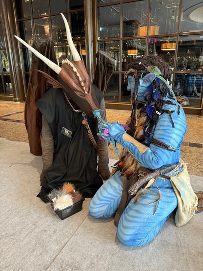 Two individuals are dressed in elaborate costumes. One features a dragon with horns and wings, while the other is adorned in blue attire with long, braided hair. They are engaged in a gesture, possibly in a playful interaction.