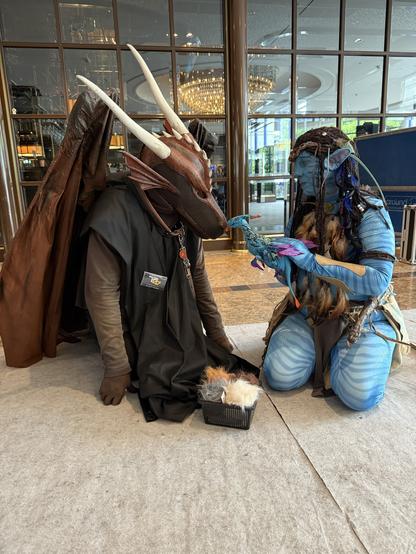 Two individuals in elaborate costumes are interacting in a lobby. One is dressed as a dragon-themed character with a mask and wings, while the other is in blue body paint resembling an avatar-like creature. They are holding a colorful, bird-like figure.
