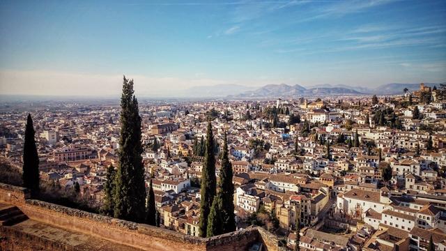 A panoramic view of Granada, Spain, showcasing a dense urban landscape of white and terracotta-roofed buildings stretching towards distant mountains under a clear blue sky. In the foreground, tall, slender cypress trees stand on a terracotta-coloured wall, providing a natural frame for the expansive city below.