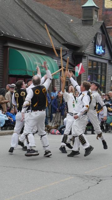 Six morris dancers with white shirts and trousers and white and black vests decorated with yellow and brown ribbon rosettes dancing in a street. They are in a tight circle, tossing their sticks into the air. An audience watches from behind.