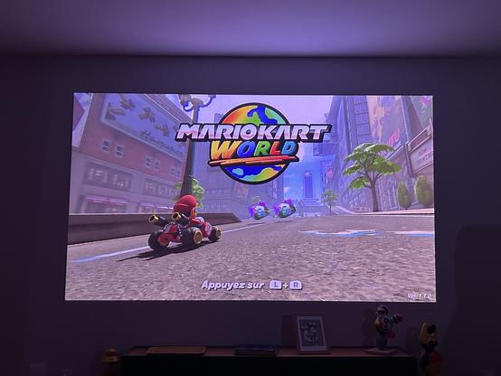 Photo de mon salon où est projeté une image de 2,90m de base. On voit l’écran d’accueil de Mario Kart World tourner. L’image est très violette car mon vidéo projecteur est laser, lorsque l’on prend une photo, les couleurs ne sont pas représentative de ce qu’observe l’oeil humain.