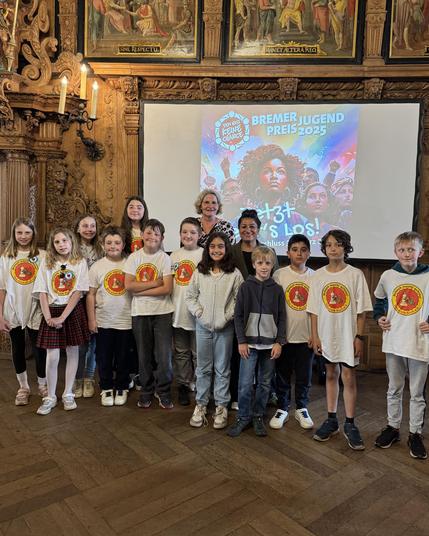 Gruppe von Kindern in weißen T-Shirts mit einem Logo, die in einem historischen Raum vor einem Bildschirm mit der Aufschrift 'Bremer Jugendpreis 2025' posieren. Sie stehen zusammen mit der Vizepräsidentin der Bremischen Bürgerschaft, Sahhanim Görgü-Philipp.