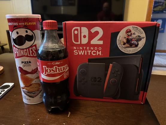 Switch 2 box sitting on wooden table with a can of pizza flavored pringles and a bottle of coke.