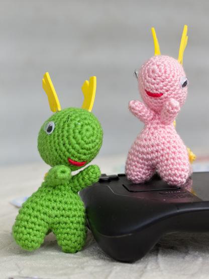 Chrocheted KDE mascotts Konqi and Katie next to a Steam Deck. Konqi, a green dragon, is left to it and Katie, a pink dragon, is on top of the gaming console. The background is blurry.