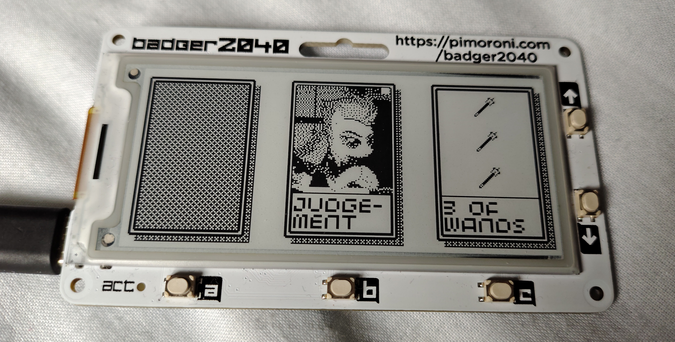 A photo of the Badger 2040 e-ink conbadge with a USB-C cable plugged into it. The e-ink display shows three representations of tarot cards. The first card is face-down. The second card is face-up, and shows the Judgement card, which depicts the character Spitfire from My Little Pony: Friendship is Magic. The third card is a minor arcana card, the Three of Wands, which is illustrated with an arrangement of three wand symbols.
