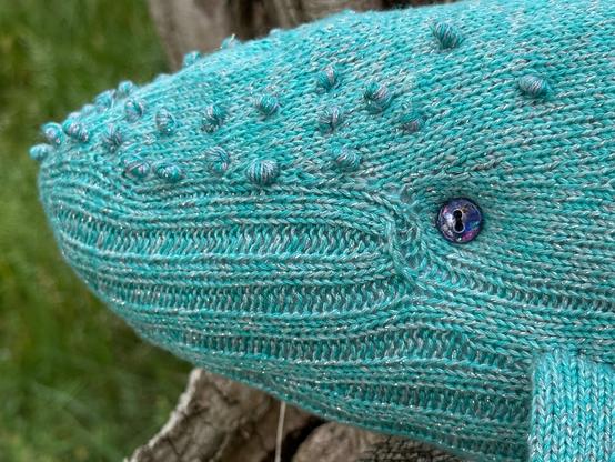 Nahaufnahme des gestrickten Buckelwal-Kopfes. Man sieht die silbrig glänzenden Fäden in der hellblauen Wolle und ein regenbogen-violett schillerndes Auge (ein aufgestickter Knopf).