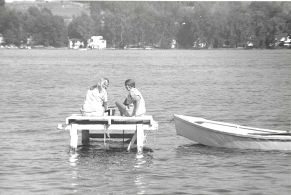 Two girls sit on a small dock over a calm lake, engaged in conversation. A rowboat is tethered nearby. Trees line the distant shore, evoking a tranquil scene.