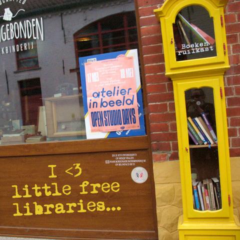 An old clock painted yellow and repurposed as a free little library in front of a house with a bookbinding workplace. Text says: "I heart little free libraries".