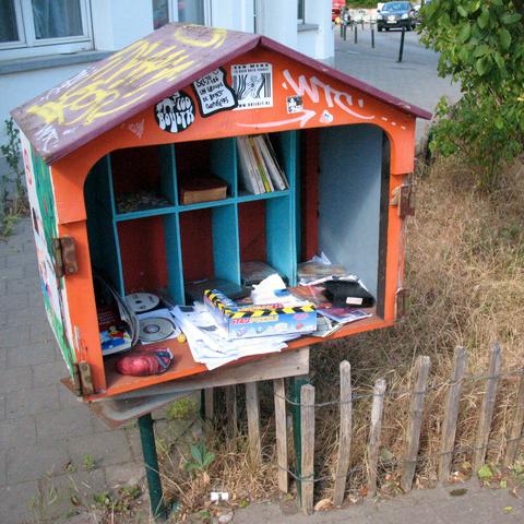 A, orange and blue little free library in the shape of a little house on the pavement. It looks messy with CDs, trash, but also some books in its shelves.