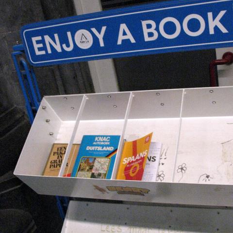 White shelves with 3 books and lots of empty space and the blue sign "enjoy a book" on top of it. It's inside a train station. The same little free library design can be found in lots of train station halls in Belgium.