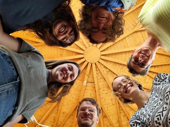 6 Personen stehen im Kreis und lachen von oben in eine Kamera. Über den Personen in deren Kreismitte ist das Holzdach der Rotunde der DNB Leipzig zu sehen; es sieht aus wie eine Sonne mit Strahlen.