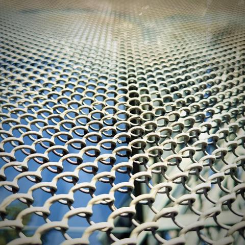 A macro perspective view of a chain-link fence shows the links extending up past the top of the frame into the distance on a sunny day.
