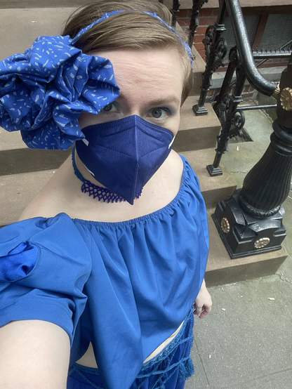 Enby in eclectic royal blue costume and matching mask taking a selfie in front of a Brooklyn brownstone