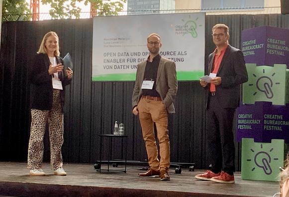 Drei Vortragende stehen auf einer Outdoor-Bühne beim Creative Bureaucracy Festival. Links steht Luisa Lamm (OKF/bydata) mit einem Tablet, in der Mitte Maximilian Melle (OKF/bydata) und rechts Olaf Neumann (Digitalagentur Brandenburg). Im Hintergrund ein Bildschirm mit dem Vortragstitel: „Open Data und Open Source als Enabler für die Nachnutzung von Daten und Systemen“. Daneben das Festival-Logo und Bühnen-Deko mit dem Icon einer Büroklammer als Glühbirne.