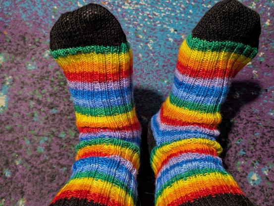 Photo of two feet in rainbow hand knit socks against a background of nebula pattern carpet.