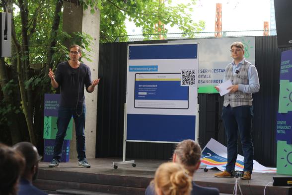 2 men on a small stage with a board between them with a pinned poster showing a screenshot of the new Service Standard website with a QR code; stacked cubes next to them with the logo of the Creative Bureaucracy Festival