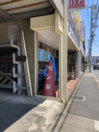 A narrow street scene featuring a storefront with a large red traffic cone displaying Japanese characters. The entrance shows shelves with blue items, and metal pipes are stacked nearby. The street is clear, with power lines visible in the background.