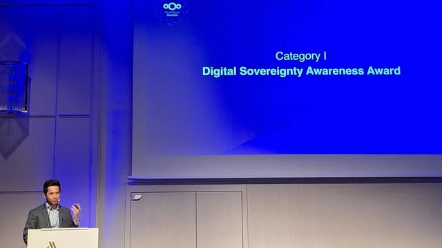Das Bild zeigt eine Veranstaltung in einem großen Saal, bei der ein Mann an einem Podium steht und spricht. Der Hintergrund ist ein großer blauer Bildschirm, auf dem in weißer Schrift "Category I Digital Sovereignty Awareness Award" steht. Der Mann trägt einen dunklen Anzug und eine hellblaue Krawatte und gestikuliert mit seiner rechten Hand. Der Podium hat ein Logo, das nicht vollständig sichtbar ist. Die Beleuchtung ist gedämpft, mit einem blauen Licht, das den Raum um den Bildschirm beleuchtet. Die Atmosphäre wirkt formell und professionell.