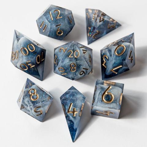 Ein Würfelset in blau mit weißen Swirls im inneren. Das Set besteht aus einem D4-Shard, einer D4-Pyramid, D6, D8, D10, D100, D12 und D20. Das Set liegt auf weißem Untergrund mit der höchsten Seite nach oben. Die Würfel sind im Bild um den D20 angeordnet. Die Würfel sind in Goldbronze bemalt.