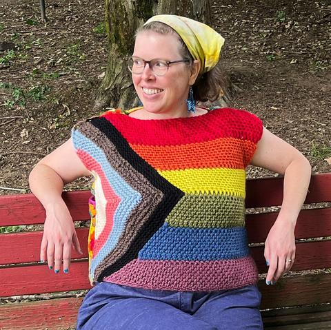 Woman, I think the designer, wearing a top with the front designed like the Pride flag with an inset triangle of white, pink, blue, gray, black stripes with rainbow colored horizontal stripes.