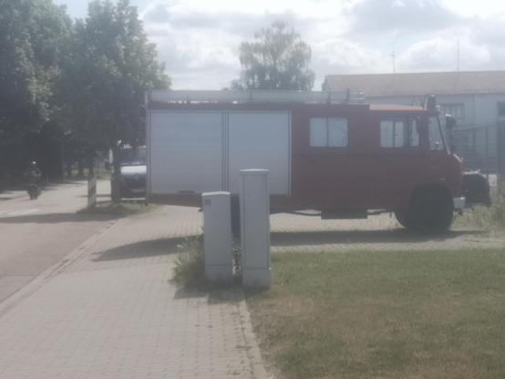 Ein altes Feuerwehrauto steht in einer Einfahrt und ragt mit dem Hinterteil um mehr als die Hälfte in den vorbei führenden Gehweg. Im Vordergrund stehen noch Schaltkästen am Rand des Gehwegs, daran grenzt ein Stück Rasen an.