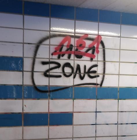 Auf einer überwiegend weiß gekachelten Wand mit blauen (gekachelten) Streifen ist ein schwarzes Graffiti
"1161
ZONE"
Der Text ist umrandet.
"1161" ist rot durchgestrichen und "161" darüber geschrieben.