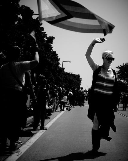"A person in sunglasses and a striped shirt walks joyfully down a street, waving their arm. They are surrounded by a crowd of people on the sidewalk. A flag waves in the foreground, capturing movement. The setting is urban, with trees lining the road.