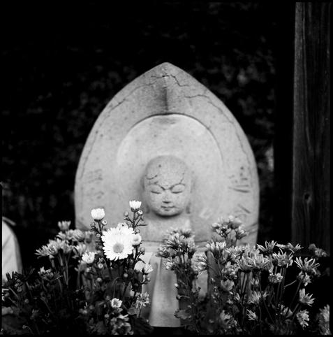black and white photograph of grave flowers in front of a small buddhist statue. the focus is on the flowers, the statue in soft focus.