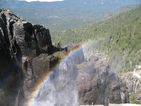 A vertical rock wall is wet with thick waterfall mist, with a rainbow of color appearing in the mist. Woods, his, and mountains are in the background.