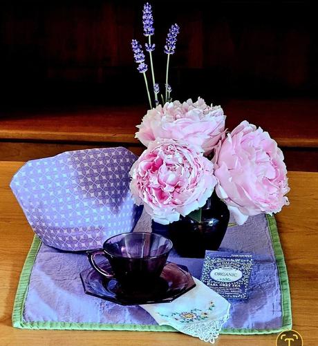 Tea tableau in purple with large pink fresh-cut peony blossoms. Placemat and cozy are purple, cup and saucer are vintage dark purple glass, and the peonies are in a dark purple vintage glass MidCentury vase. There is also an embroidered handkerchief, and a packet of organic Earl Grey tea from St Dalfour.