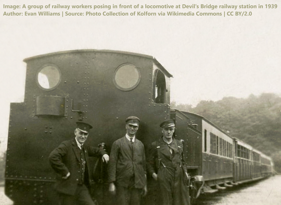 Image: An old sepia toned photo of three railway workers standing with their backs to a train and its carriages.
