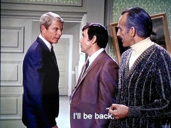 Freeze frame of a television film with three men, two of them in suits, one with an elegant cardigan from the 1960s.A subtitle says: I'll be back.
