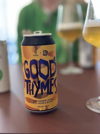 A can of “Good Thymes” fizzy summer lager—a collaboration between Nurme Brewery and Bite Me—sits on a table at Bite Me, a bar in Rīga’s Grīziņkalns district, right next to Ziedoņdārzs park. The beer is brewed with thyme and quince, and in the background, glasses of the same golden lager and a person in a green shirt add to the relaxed, summery atmosphere.