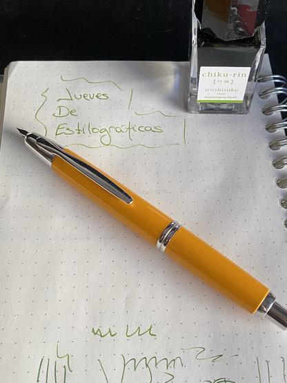 Una pluma naranja con el plumín retráctil apoyada sobre un cuaderno donde se ha escrito a mano “Jueves de estilográficas”. Al lado del texto se ve un pequeño bote de cristal con tinta verde.
