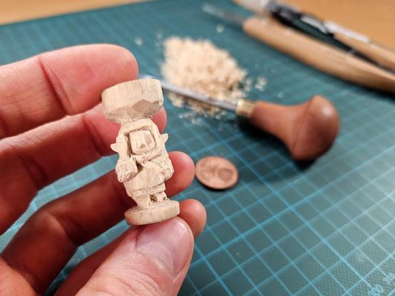 Foto einer Hand, die eine kleine Holzfigur hält. Dahinter eine grüne Schneidenatte auf der Holzspähne und Schneidewerkzeuge liegen.