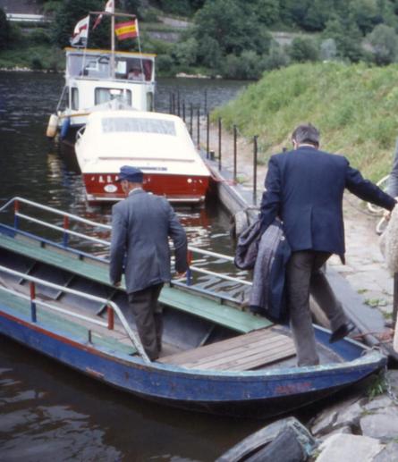 Die Ruderfähre für ca. 10 Personen liegt am Ufer, der Fährmann und eine weitere Person betreten das Boot. Die Fähre hat rechts und links Holzbänke mit einer Metallrückenlehne und am Heck eine einfache kurze Bank.