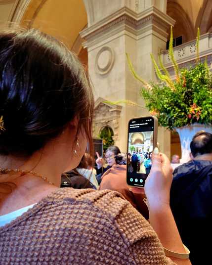 A woman in a brown knit sweater holds up her phone in a bustling museum hall. Onscreen, the Aira call reads “Connected with Agent Julie,” displaying the grand lobby and crowd. Floral arrangements and arches rise behind.