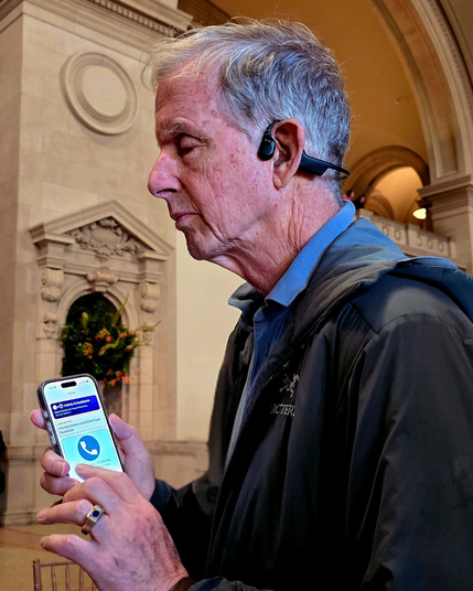 An older man in a dark jacket and bone-conduction headset holds his phone, using the Aira app. The screen shows the Aira home page with the button to “Call an Aira Agent.” Stone arches and floral decor rise behind him.