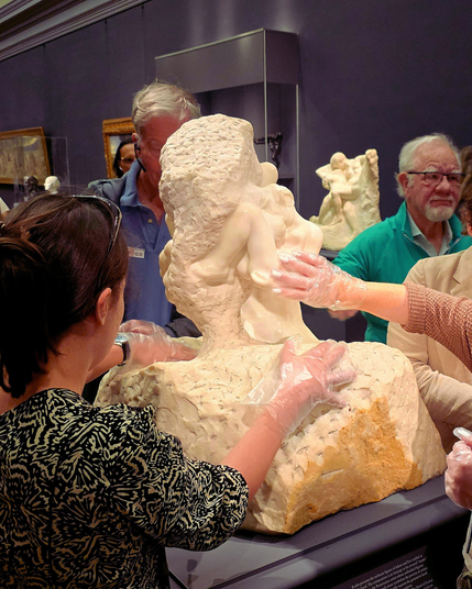 Visitors wearing gloves explore a textured marble sculpture of intertwined figures by touch. A man in blue and a woman in a printed dress guide the interaction. Framed artwork and another sculpture are visible in the gallery background.