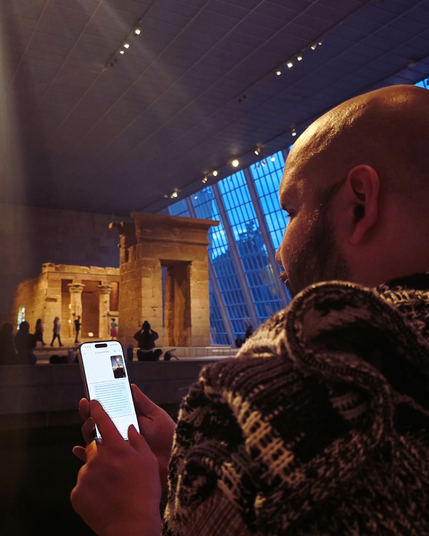 A man in a patterned sweater uses Aira AI at the Temple of Dendur exhibit at the Met. His phone displays the Aira app with a text chat. Evening light filters through the vast glass wall behind the temple.