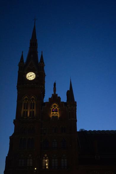 Against a darkening deep blue sky stands the enshadowed tower of St Pancras hotel, topped by a steeple flanked by shorter spikes and neighboured by a steep-stepped gable end, its own sharp-topped turret and a lower roof. In the tower glows a white glass clock, divided by black leading and faced with roman numerals; below, high-arched tall windows are lit from beneath, standing out against the void of the silhouetted building.