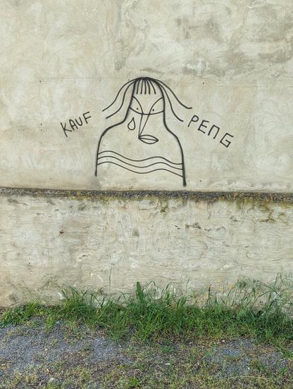 A raw, unedited, eye-level, full shot shows a graffiti artwork on a textured, light-grey concrete wall. The central feature is a stylised drawing of a human figure with a teardrop falling from its eye. To the left of the figure, the word 'KAUF' is spray-painted, and to the right, 'PENG'. The wall has a darker, horizontal line running across its lower section. Below the wall, a strip of green grass grows over a gravel patch.