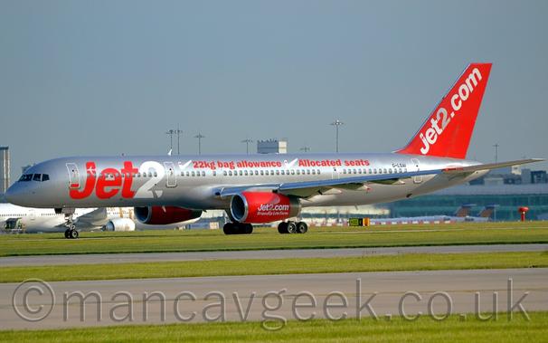 Side view of a twin engined jet airliner moving from right to left.
The plane is mostly a shiny silvery-grey, with red "jet2" billboard-stye titles (the "2" in white) on the forward fuselage, with additional red "22kg bag allowance" and "Allocated Seats" text next to large white check marks on the upper fuselage.
There are large white and grey "Jet2[dot]com" titles on the bright red tail, as well as on the engine pods mounted under the wings.
The engines also have smaller, white "Friendly low fares" in an almost hand-written font.
Taxiways lined with vivid green grass stretch across the foreground, while a grey terminal building stretches across the background, with several other planes visible parked in front, all under blue-grey sky.