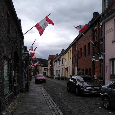 In einer gepflasterten Straße parken einige PKW. An den Häusern links und rechts der Straße hängen rot-weiss-rote Fahnen mit einem Wappen. Der Himnel ist bewölkt.