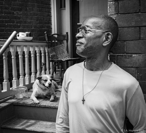Black and white photograph of a man leaning against a brick wall by the entrance to an apartment. A white dog is sitting on the landing above the stairs.