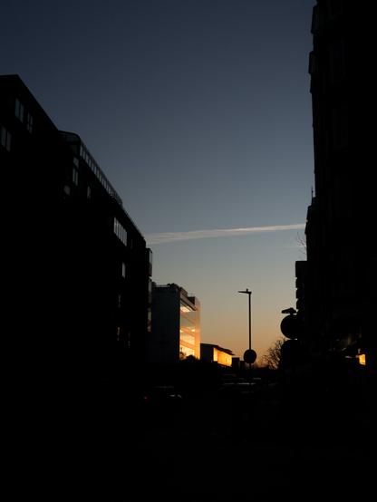Buildings run down both sides of a wide street mostly silhouetted under a faded indigo denim sky that reflects in their windows. The orange flame of sunset lights up a couple at the far end.