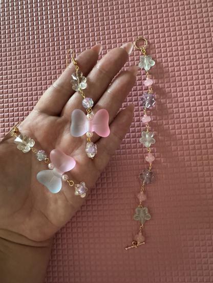 a set of wire earrings and a wire bracelet. the earrings have big bow beads. the bows are transparent pink and purple split in the middle, i surrounded them with small pink fake pearls and hung star charms from the bottom. on top i used a sphere bead that’s transparent and sort of a pink/purple mix and above that is a clear bear bead. the bracelet is a wire wrapped pattern of a transparent seafoam flower, then a pink seed bead pink i think quartz? idr a pink flattened rock bead it’s like a rounded rock bagel shape. then another pink seed bead. then an extra transparent blue flower, then another seed bead/rock bead, and back to the seafoam flower.