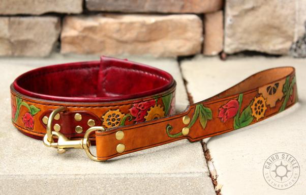 A handmade leather collar with attached short leash. The collar and leash have medium brown base straps with painted tooling depicting green vines with red flowers intertwining golden gears. The collar has dark brown top straps and an oxblood liner, with dark brown stitching. Both pieces have brass hardware.