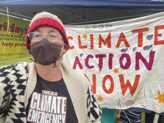 Woman wearing beanie and mask and a Climate Emergency tshirt, stands in front of Climate Action Now sign